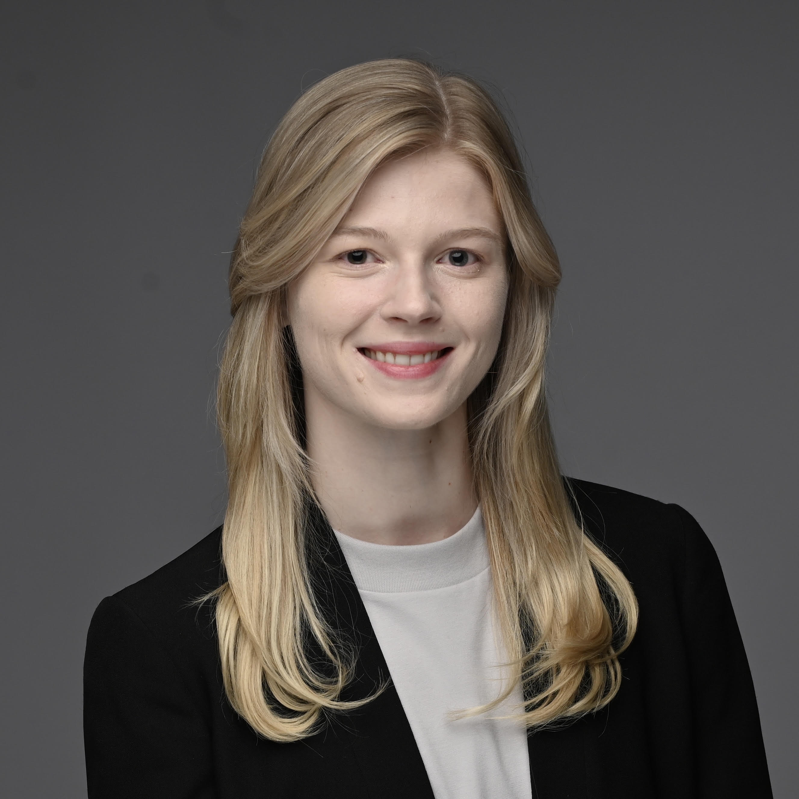 A studio headshot of Erin. They are smiling, have medium-long blond hair and are wearing a black blazer over a white top, set against a dark grey studio background.