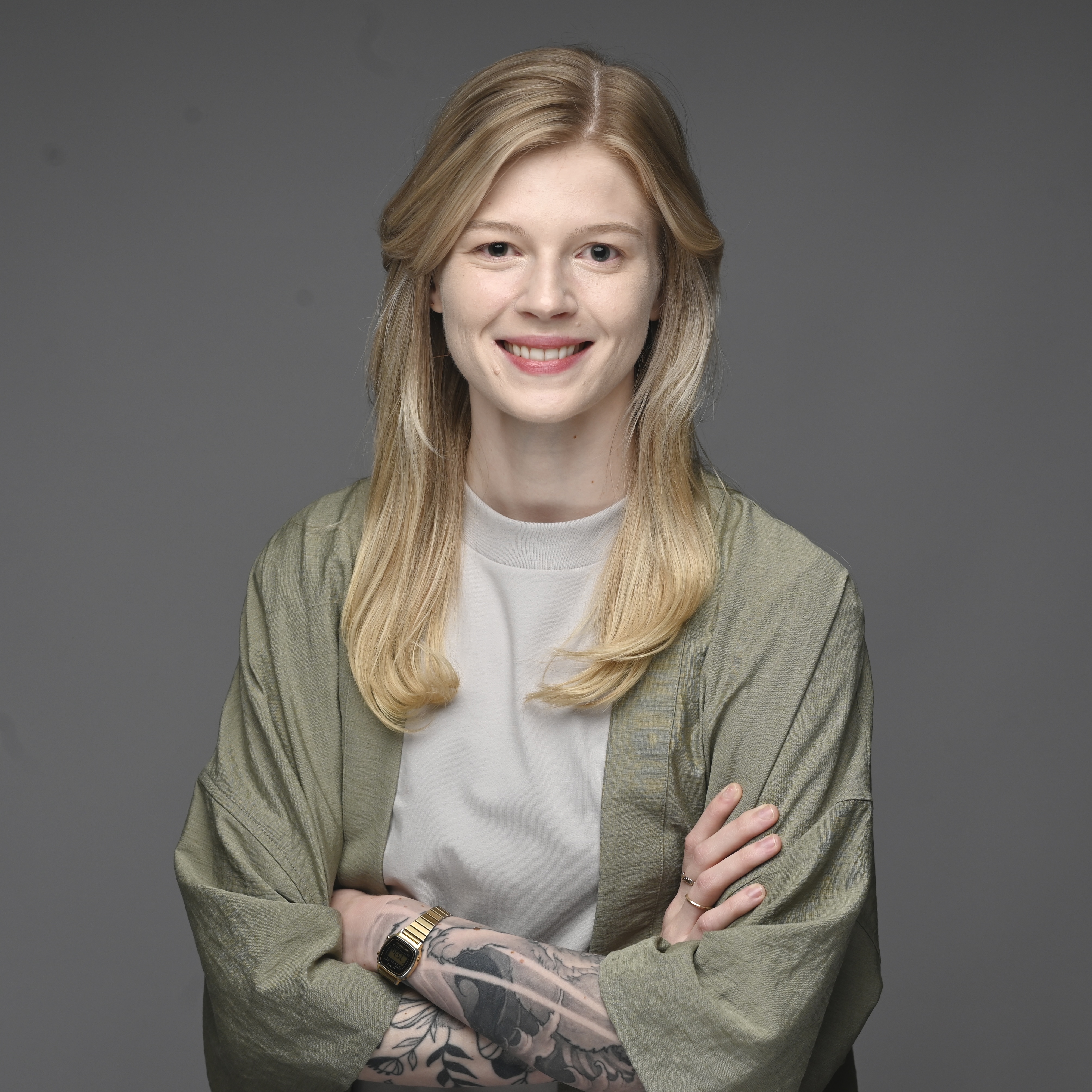 A studio headshot of Erin, a smiling white person with long blonde hair, standing against a dark gray background with their arms crossed. They are wearing a light green jacket over a light gray tshirt, with visible forearm tattoos, gold rings, and a bronze digital watch.