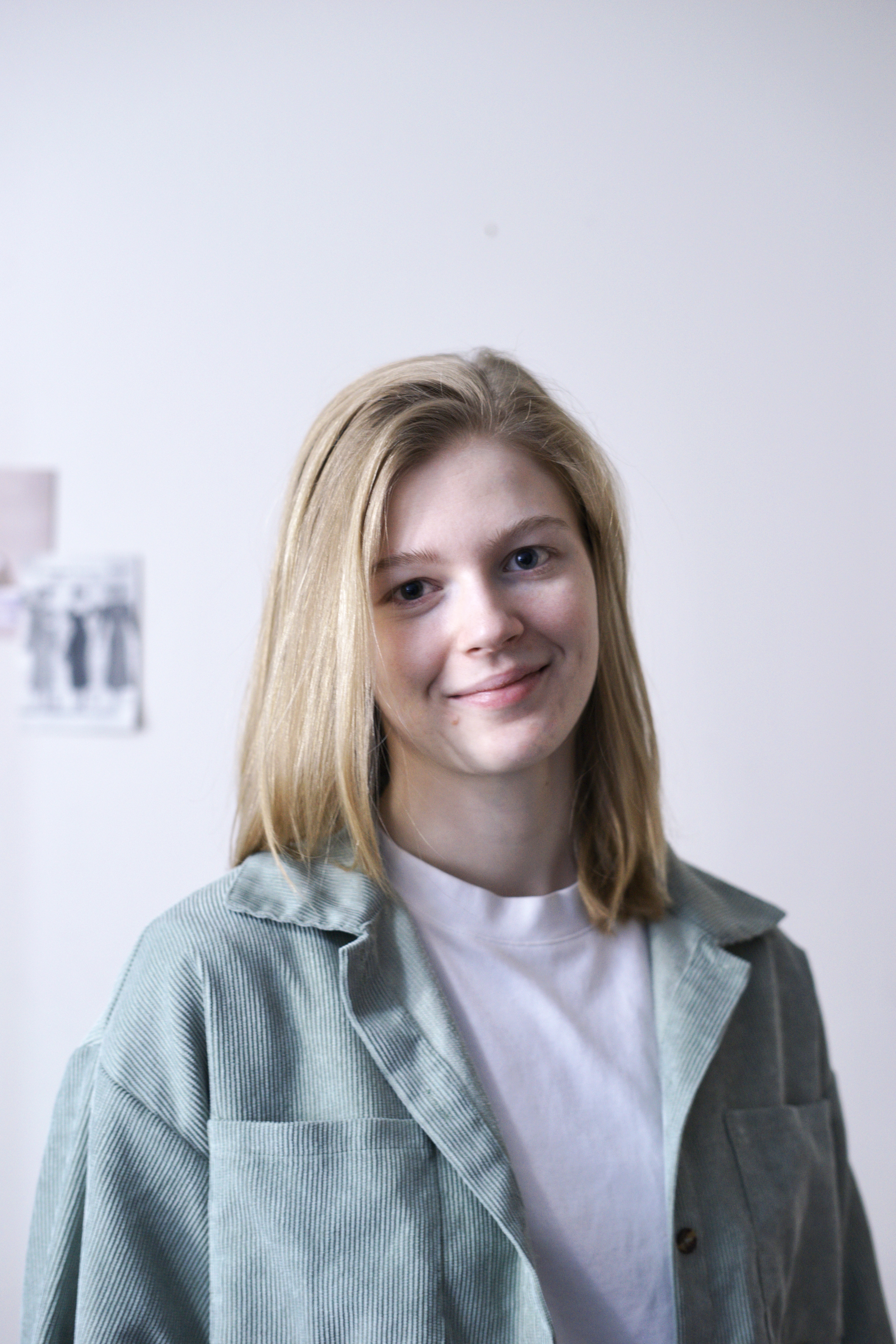 Erin Dekker has shoulder-length blonde hair, and is wearing a light green corduroy jacket over a white t-shirt. The background is softly lit, with a neutral white wall with blurred artwork pinned in the upper left corner of the background.